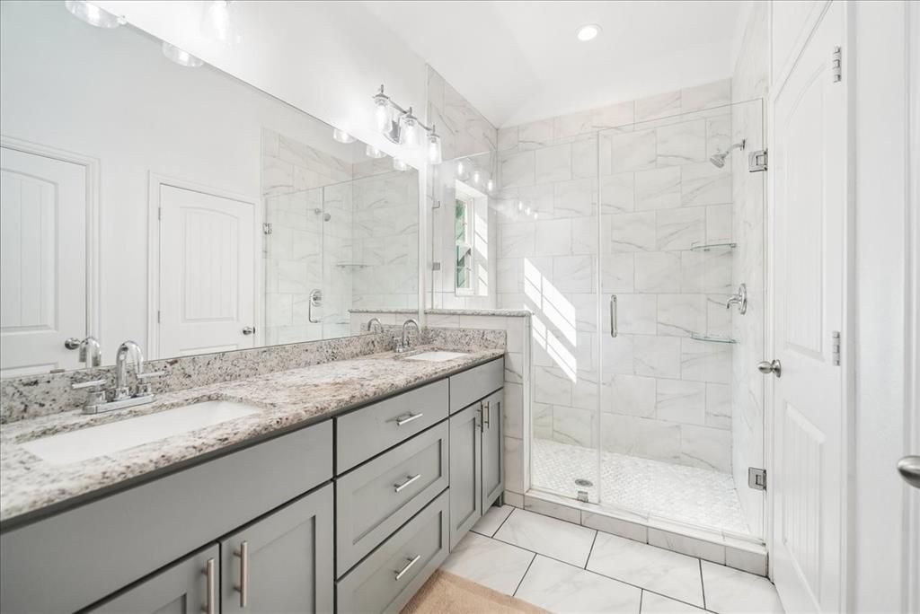 Bathroom featuring a grey dual-sink vanity with a stone countertop, a glass-enclosed shower with tiled walls and flooring, and full-width vanity mirror with integrated lighting