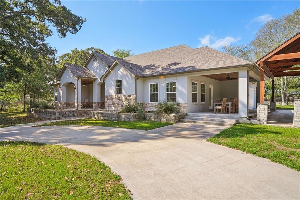 Single-story exterior featuring a light-colored masonry facade with stone veneer accents, a concrete driveway, a covered patio area with a wood-plank ceiling, and a detached wooden pergola structure