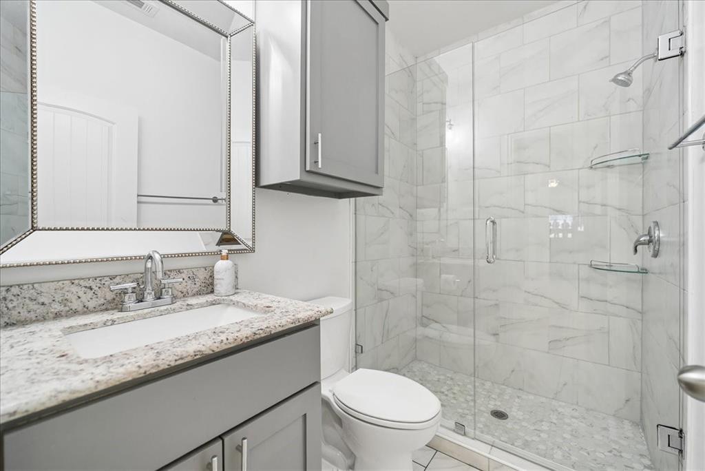 Bathroom featuring a granite vanity countertop, gray shaker-style cabinetry, a framed wall mirror, a glass-enclosed shower with stone-tile surround, and a pebble-stone shower floor