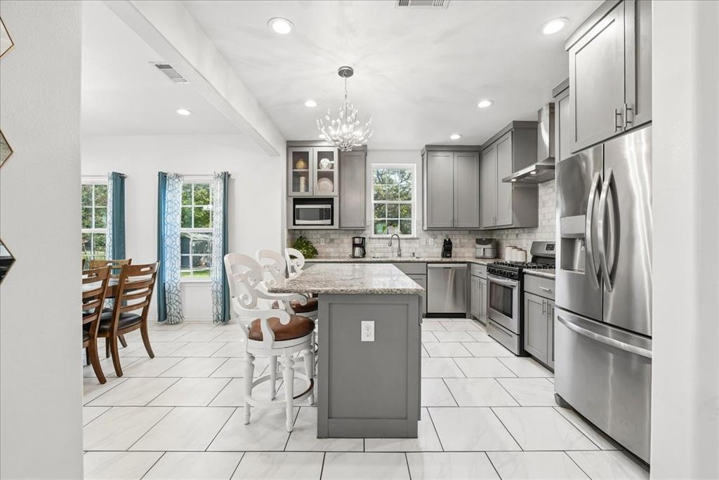 Kitchen featuring gray shaker-style cabinetry, a central island with stone-surface countertop, stainless steel appliances, a light-toned tile backsplash, and decorative chandelier lighting