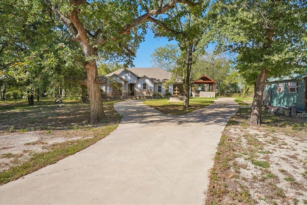 Single-story residence featuring a bifurcated concrete driveway, stone and neutral-tone siding exterior, decorative water feature, and a detached wooden-frame outdoor pavilion situated on a lot with mature trees