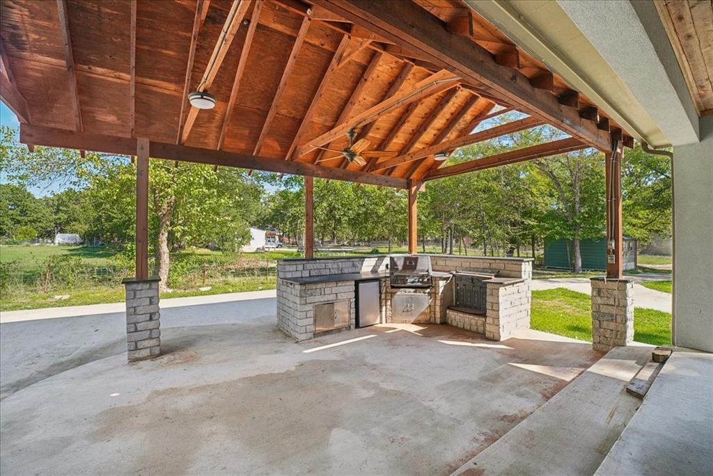 Covered outdoor kitchen featuring a timber-framed vaulted ceiling, stone pillar supports, stainless steel built-in grill, integrated refrigeration, and masonry countertop