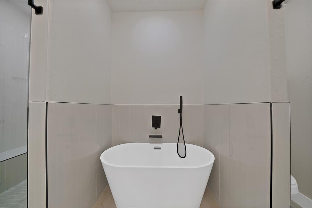 Freestanding white soaking tub with matte black wall-mounted faucet and handheld shower sprayer, complemented by floor-to-ceiling neutral tile surround