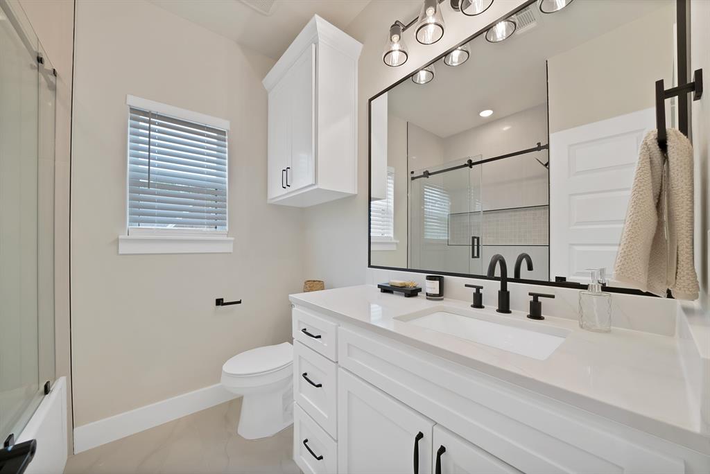 White vanity with quartz countertop, matte black widespread faucet, framed vanity mirror, overhead industrial-style lighting, and built-in upper storage cabinet