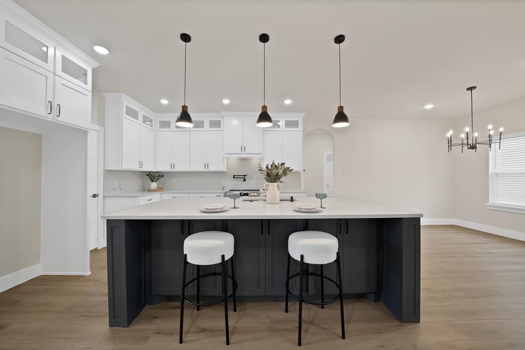 Kitchen featuring white cabinetry with glass-paneled uppers, central island with dark finish and light-toned countertop, wood-finish flooring, and recessed lighting with pendant fixtures