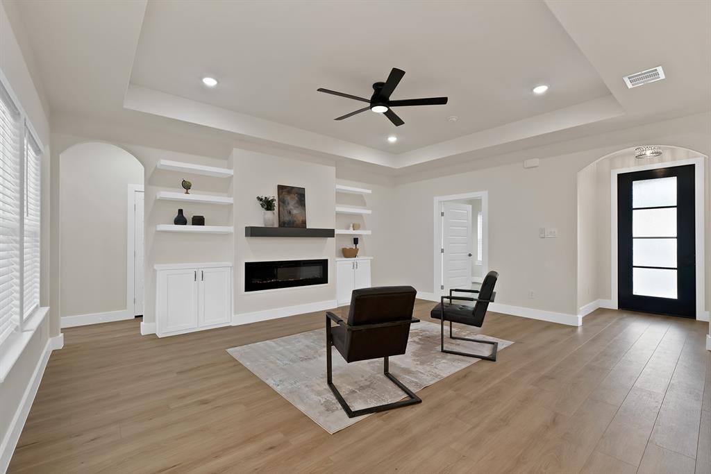 Tray ceiling, linear electric fireplace with custom mantle, built-in cabinetry with floating shelving, wood-finish flooring, and black-framed glass door