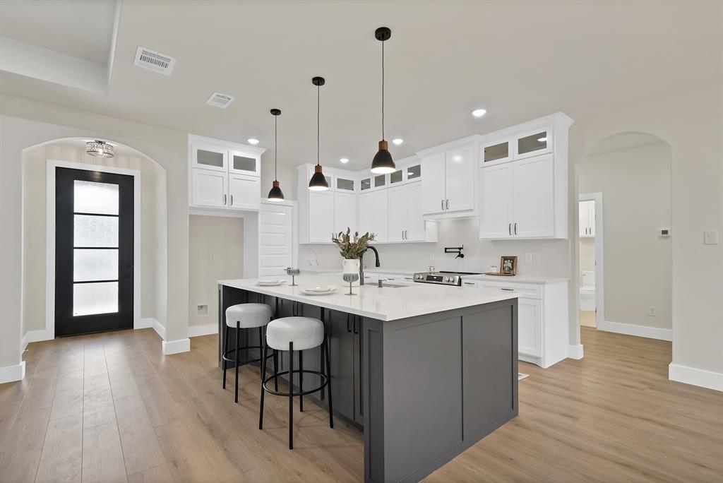 Kitchen featuring a central island with a contrasting dark base, white cabinetry with glass-front uppers, light-toned wood-finish flooring, quartz countertops, and three minimalist pendant lights