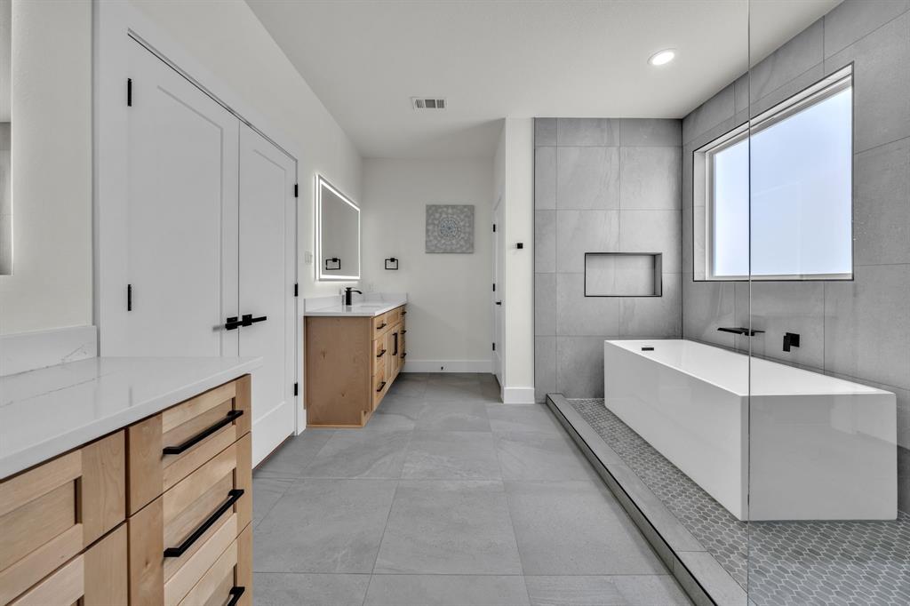 Bathroom featuring a freestanding rectangular soaking tub, large-format grey floor and wall tile, dual wood-finish cabinetry with white stone countertops, and matte black hardware