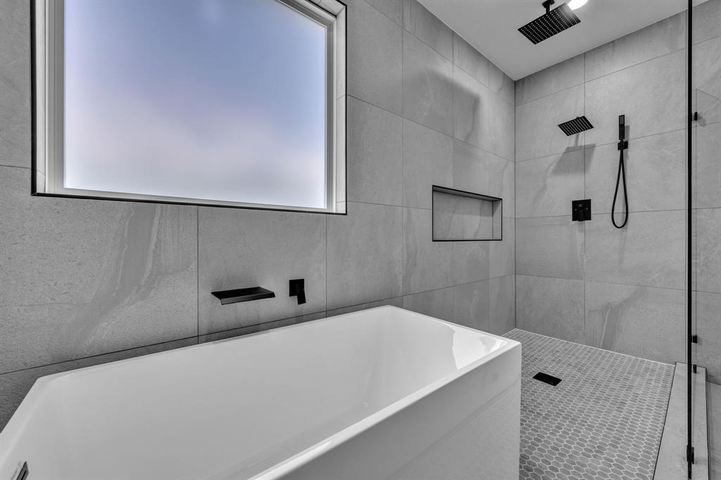 Freestanding soaking tub with matte black wall-mounted faucet, walk-in shower featuring large-format tile walls, ceiling-mounted rainfall showerhead, handheld shower fixture, and built-in wall niche