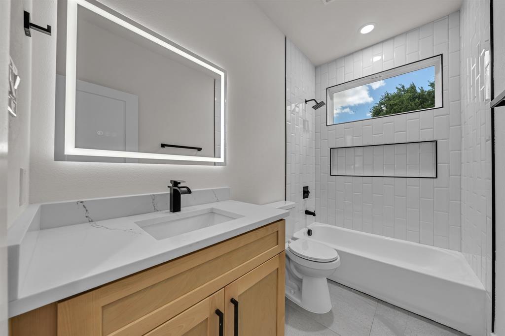 Bathroom featuring a vanity with wood-finish cabinetry, white countertop, integrated rectangular sink, and matte black faucet, complemented by an LED-backlit mirror, subway-tiled tub surround with a recessed niche, and horizontal transom window