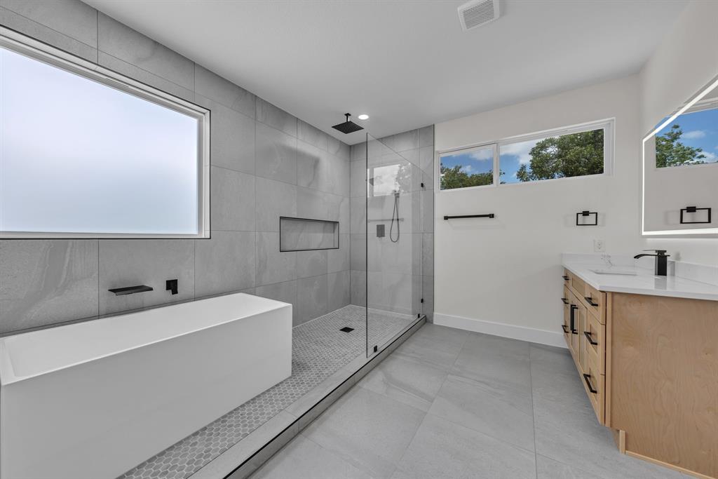Bathroom featuring a freestanding soaking tub, walk-in glass-enclosed shower with a built-in niche, light wood-finish vanity with a stone countertop, large-format floor tiling, and matte black fixtures