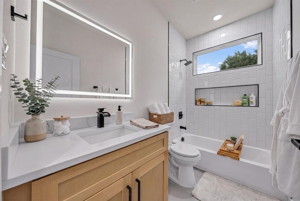 Light-toned wood vanity with a white countertop, rectangular undermount sink, matte black faucet, and rectangular LED-backlit mirror