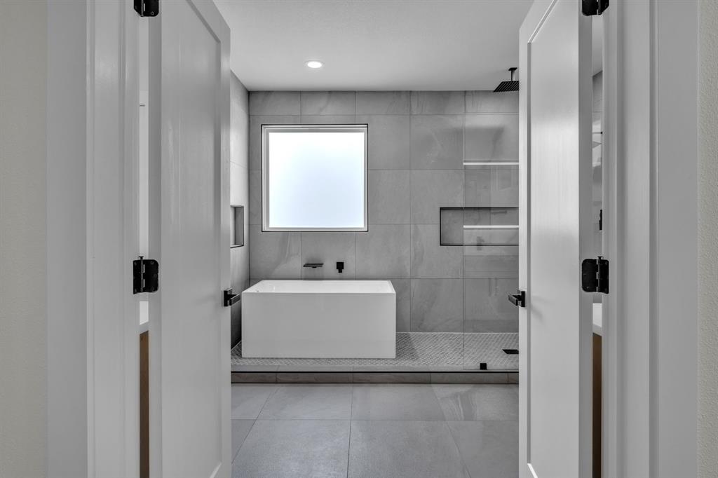 Freestanding soaking tub, walk-in shower with glass enclosure, floor-to-ceiling large-format wall tile, built-in recessed wall niches, and matte black fixtures