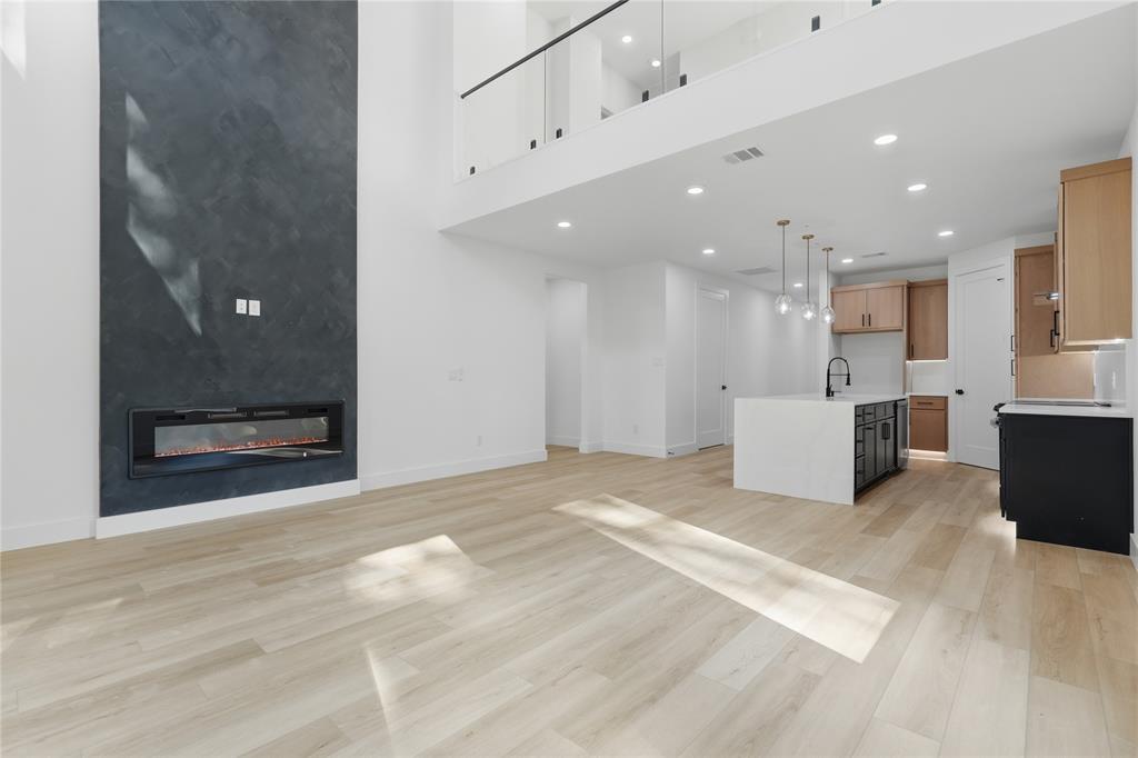 Double-height living area featuring a floor-to-ceiling dark textured fireplace surround with a linear electric insert, wood-finish flooring, recessed lighting, and a glass-paneled mezzanine railing
