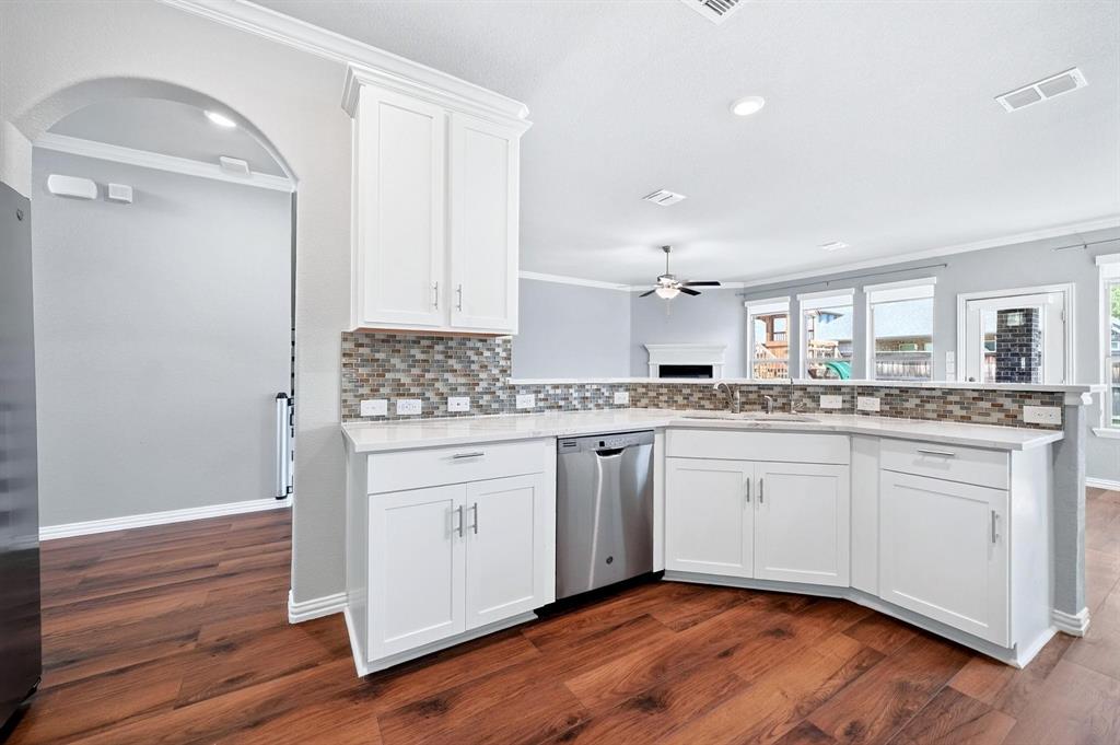 Kitchen featuring white shaker-style cabinetry, a multi-tone tile backsplash, stainless steel dishwasher, stone-finish countertops, and wood-finish flooring