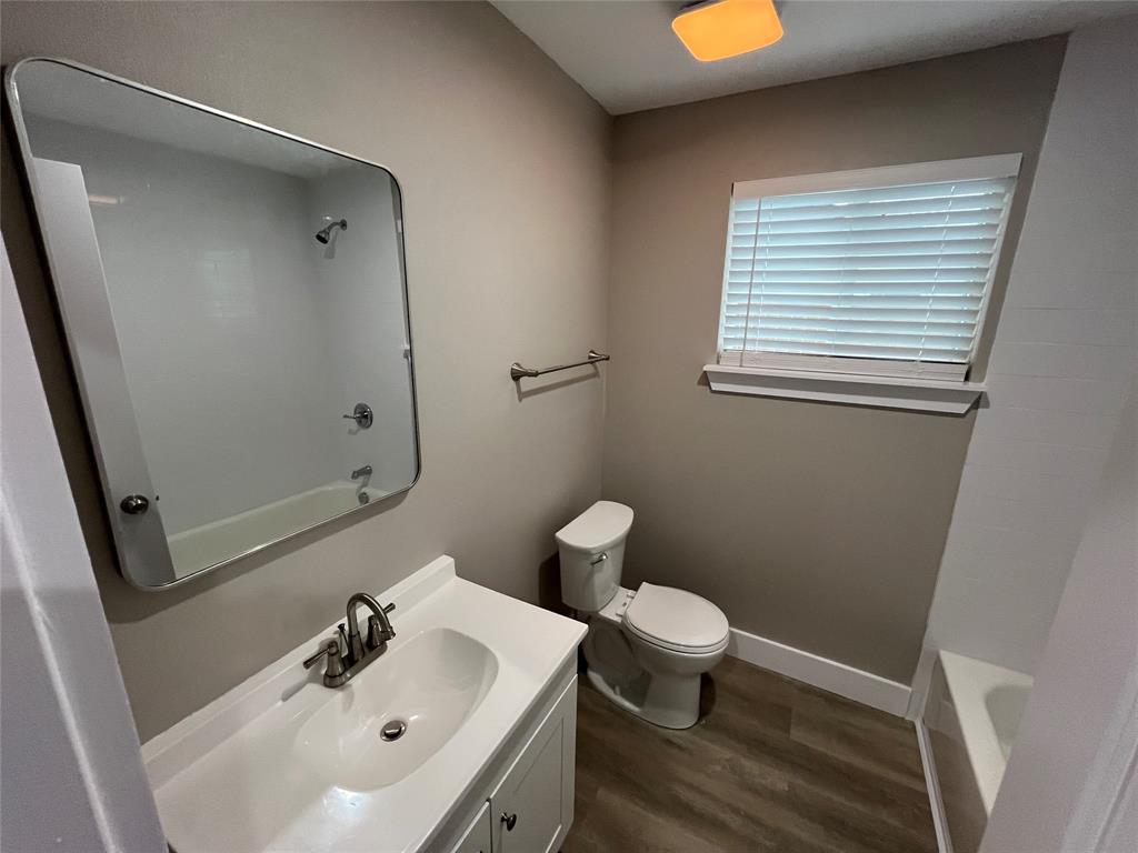 Bathroom featuring wood-finish flooring, a white vanity with an integrated sink and metal faucet, a large rectangular mirror with rounded corners, and a bathtub with a shower fixture