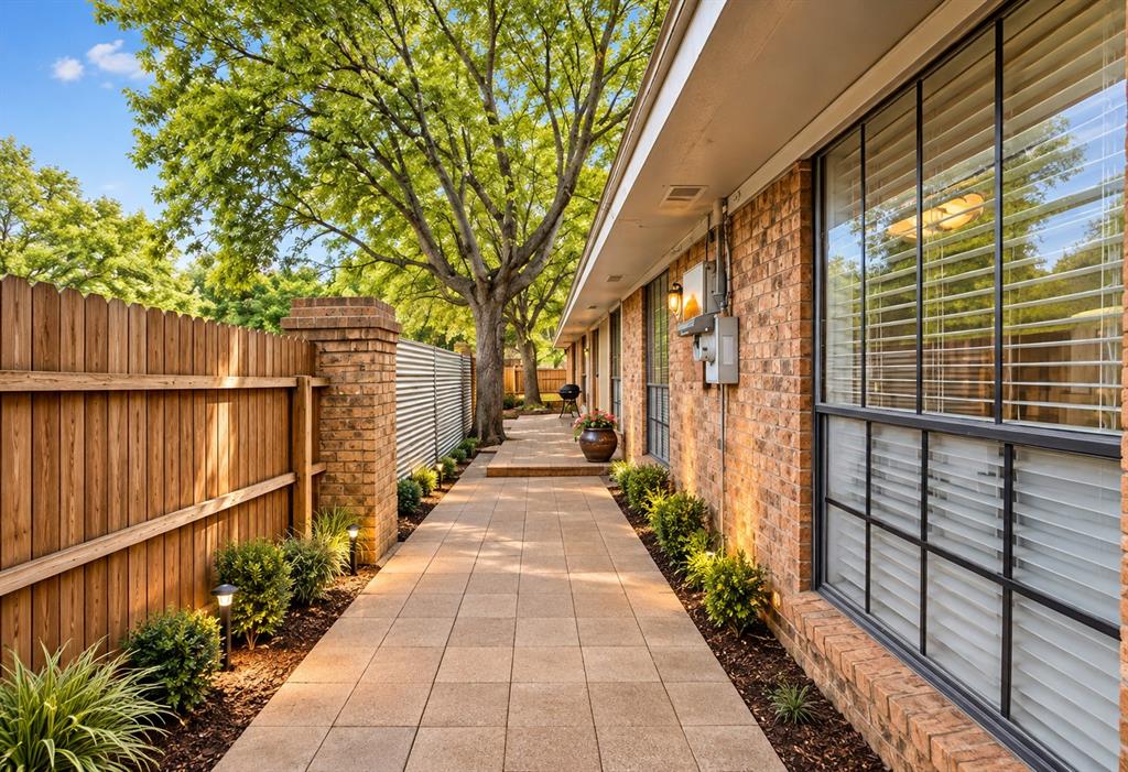 Side exterior featuring a brick facade, paved stone walkway, wood privacy fencing, integrated low-voltage lighting, and mature trees