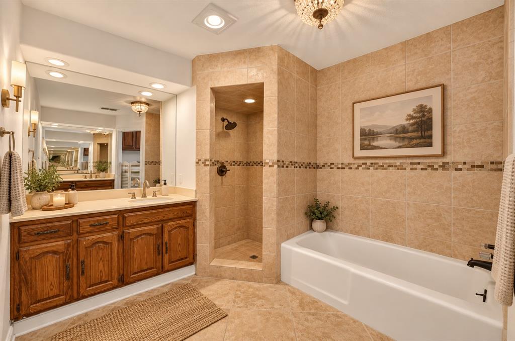 Bathroom featuring a custom wood-finish vanity with a countertop, recessed shower with tiled walls and an accent border, soaking tub, and wall-mounted light fixtures