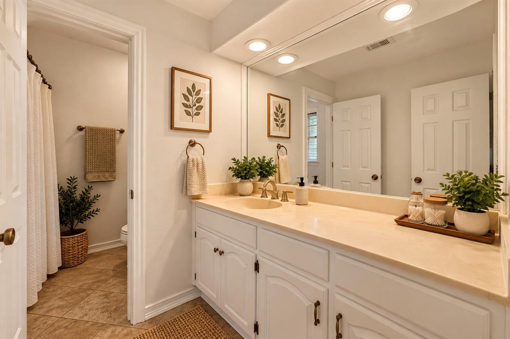 Bathroom featuring white cabinetry, solid-surface vanity countertop, inset sink with metallic faucet, full-width wall mirror with recessed lighting, and tile flooring
