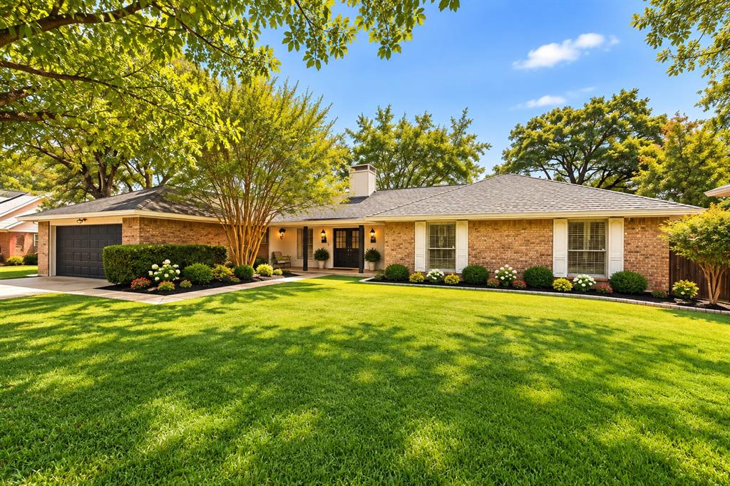 Single-story brick exterior featuring a multi-gabled shingle roof, two-car garage, manicured landscaping, and mature trees