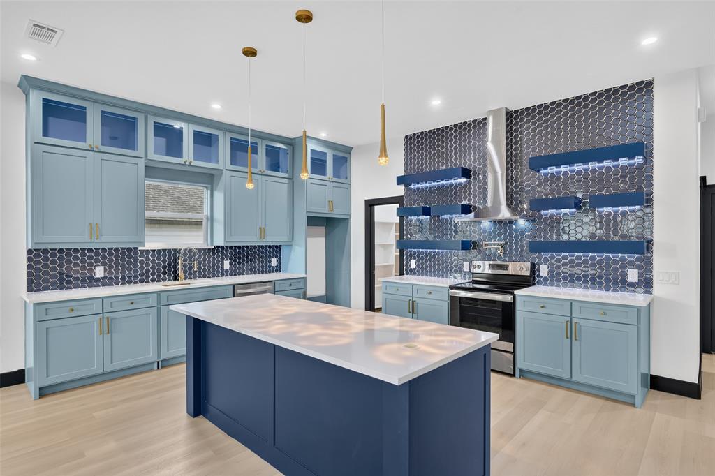 Kitchen featuring light blue cabinetry, center island with quartz countertop, hexagonal tile backsplash, stainless steel range with chimney-style hood, and tiered floating shelving with integrated lighting Kitchen featuring light blue cabinetry, center island with quartz countertop, hexagonal tile backsplash, stainless steel range with chimney-style hood, and tiered floating shelving with integrated lighting
