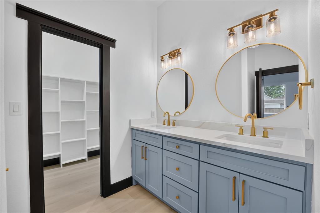 Dual-sink vanity with light blue cabinetry, gold-tone hardware, quartz countertops, round gold-framed mirrors, and brass-finish wall lighting