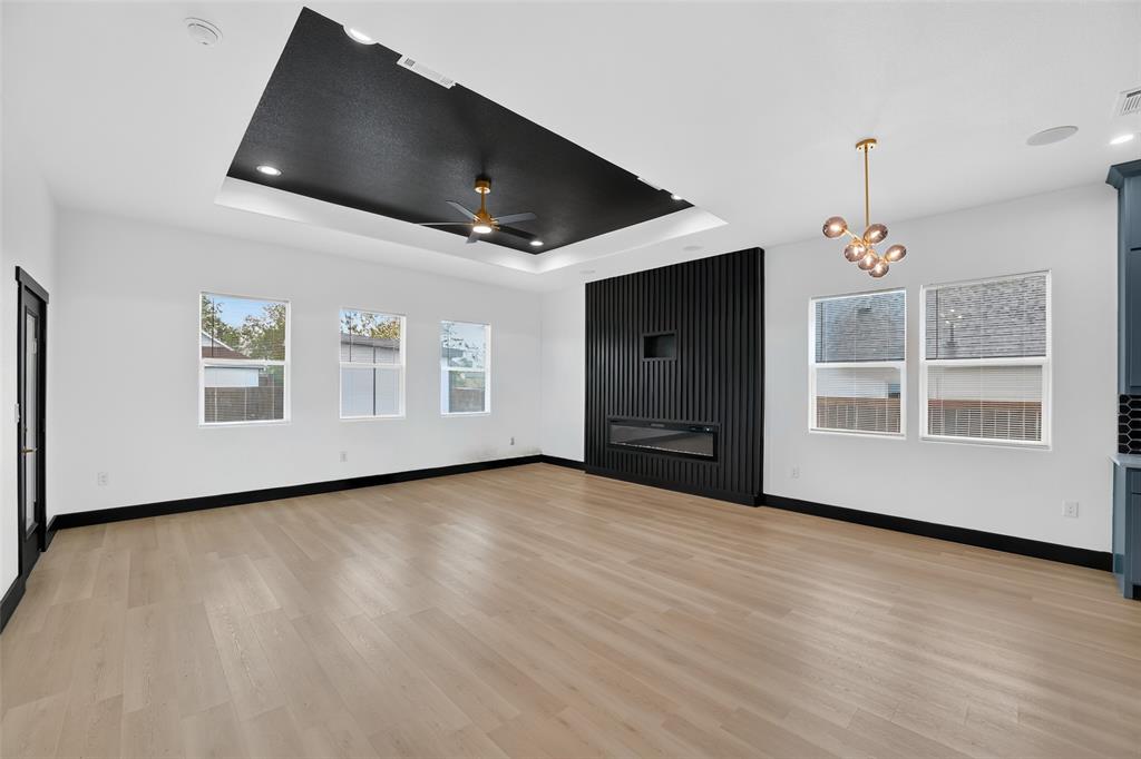 Tray ceiling with black recessed lighting, wood-finish flooring, vertical slat feature wall, built-in electric fireplace, and multi-light brass fixture Tray ceiling with black recessed lighting, wood-finish flooring, vertical slat feature wall, built-in electric fireplace, and multi-light brass fixture