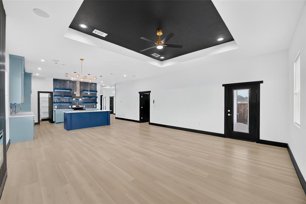 Open-concept interior featuring wood-finish flooring, a tray ceiling with a central fan, a kitchen island with contrasting cabinetry, a decorative tiled backsplash, and recessed lighting Open-concept interior featuring wood-finish flooring, a tray ceiling with a central fan, a kitchen island with contrasting cabinetry, a decorative tiled backsplash, and recessed lighting