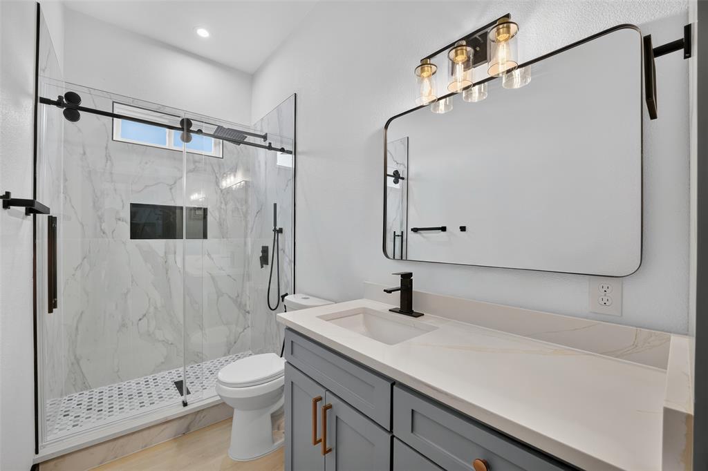 Gray cabinetry with matte hardware, stone-composite countertop, glass-enclosed walk-in shower with stone-tile surround, and rectangular wall-mounted mirror