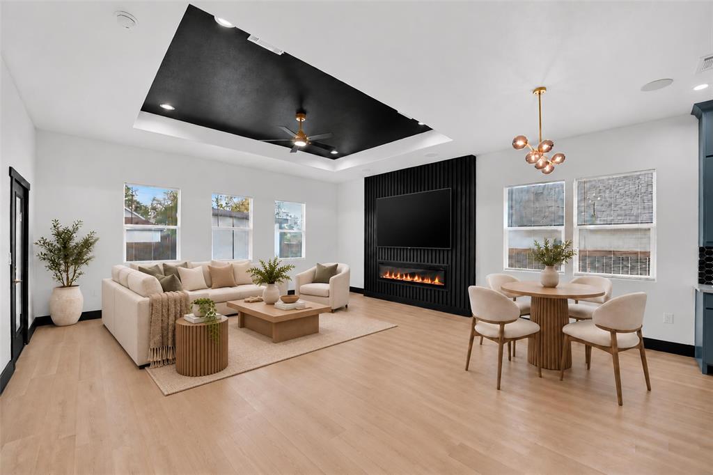 Open-concept area featuring a black recessed tray ceiling with an integrated ceiling fan, light wood-finish flooring, and a vertical-slat accent wall with a built-in linear electric fireplace Open-concept area featuring a black recessed tray ceiling with an integrated ceiling fan, light wood-finish flooring, and a vertical-slat accent wall with a built-in linear electric fireplace