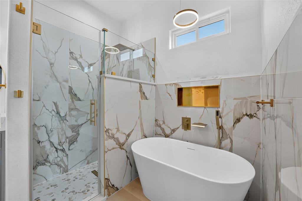 Freestanding soaking tub with gold-tone wall-mounted hardware, glass-enclosed shower with large-format stone-patterned wall tile, recessed tiled shower niche, and circular LED pendant lighting