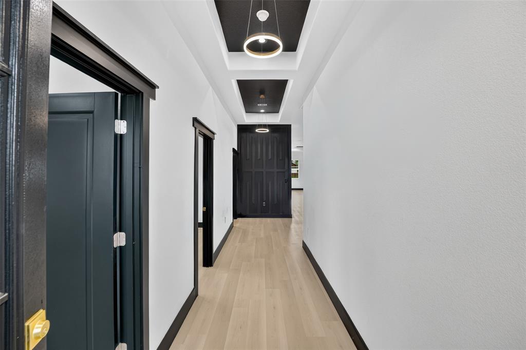 Hallway featuring wood-finish flooring, recessed tray ceilings with dark-toned architectural accents, contemporary circular pendant lighting, and high-contrast black interior door frames and trim Hallway featuring wood-finish flooring, recessed tray ceilings with dark-toned architectural accents, contemporary circular pendant lighting, and high-contrast black interior door frames and trim