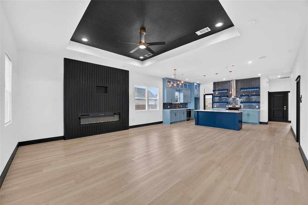 Open-concept interior featuring light wood-finish flooring, a tray ceiling with a black accent inset, a floor-to-ceiling vertical-slat fireplace surround, a central kitchen island with blue cabinetry, and a geometric-tiled backsplash Open-concept interior featuring light wood-finish flooring, a tray ceiling with a black accent inset, a floor-to-ceiling vertical-slat fireplace surround, a central kitchen island with blue cabinetry, and a geometric-tiled backsplash