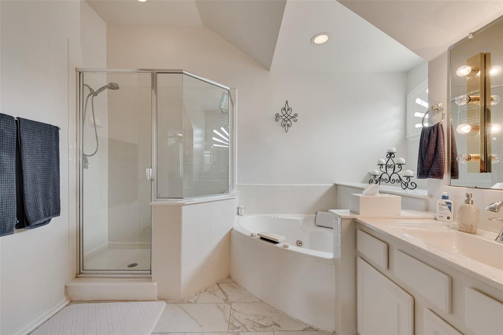 Corner soaking tub with tile surround, glass-enclosed shower, built-in vanity with integrated sink, and wall-mounted vanity lighting