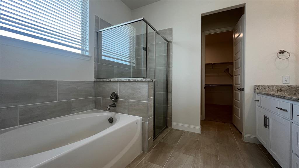 Soaking tub with horizontal tile surround, glass-enclosed shower, stone-finish vanity countertop, white shaker-style cabinetry, and tile flooring