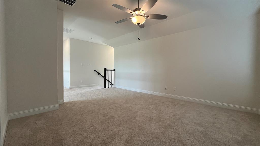 Neutral-toned wall-to-wall carpeting, vaulted ceiling with a central brushed-nickel ceiling fan, and a staircase featuring a black metal balustrade