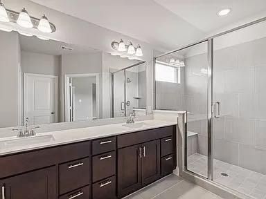Dark-finish cabinetry featuring dual undermount sinks, stone countertops, expansive wall-to-wall mirror, glass-enclosed shower with built-in bench, and tile flooring