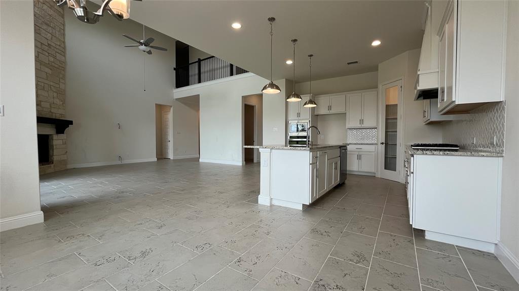 High-ceilinged interior featuring a floor-to-ceiling stone fireplace, large kitchen island with integrated sink, shaker-style cabinetry, patterned tile flooring, and recessed pendant lighting