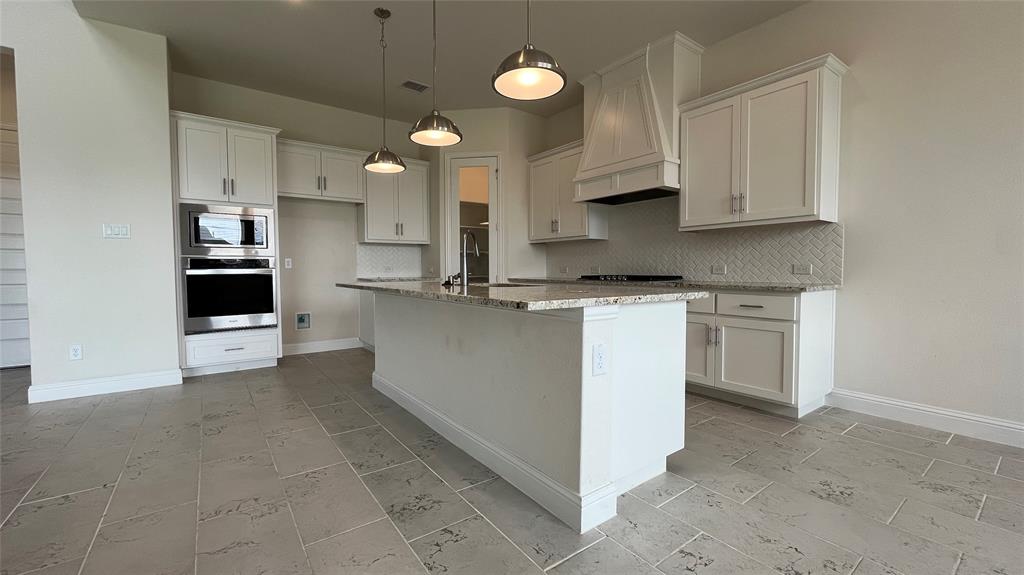 Kitchen featuring white cabinetry, stone countertops, a center island with a built-in sink, stainless steel wall-mounted oven and microwave, and herringbone tile backsplash