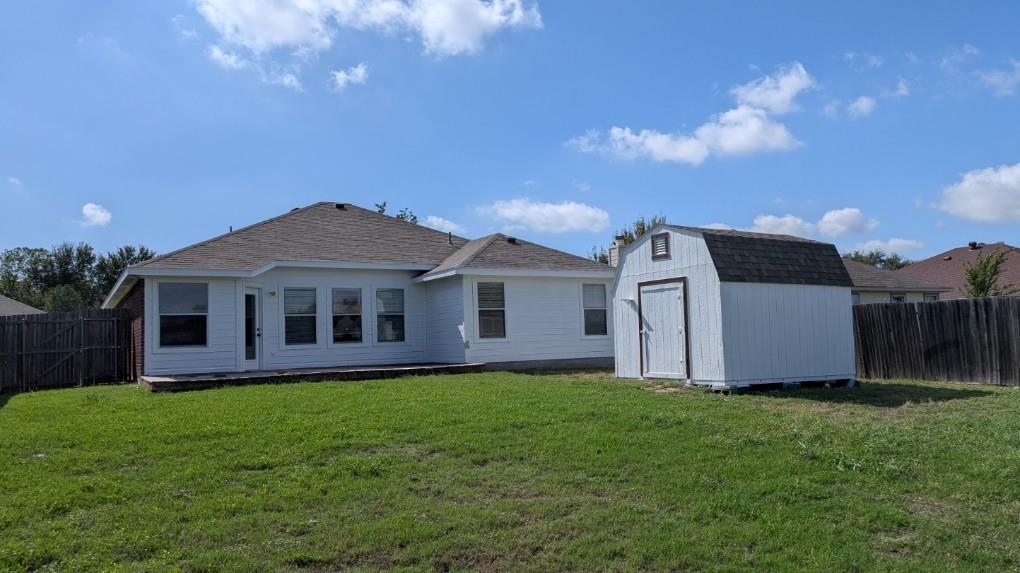 Rear exterior featuring a white siding finish, asphalt shingle roof, expansive lawn, detached storage structure, and wood privacy fencing
