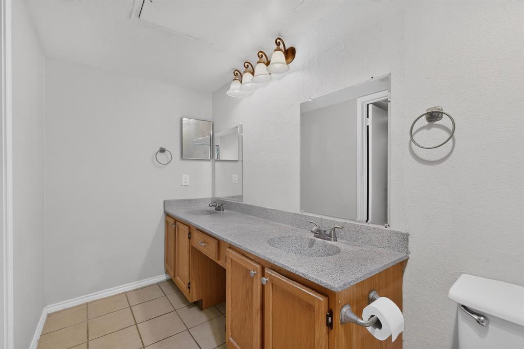 Dual sink vanity with wood-finish cabinetry, speckled countertop, and tiled flooring