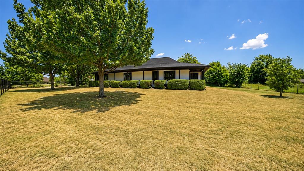 Single-story residence featuring a light-colored exterior, dark-toned roof, wrap-around covered porch, and mature shade trees situated on a sprawling lot Single-story residence featuring a light-colored exterior, dark-toned roof, wrap-around covered porch, and mature shade trees situated on a sprawling lot