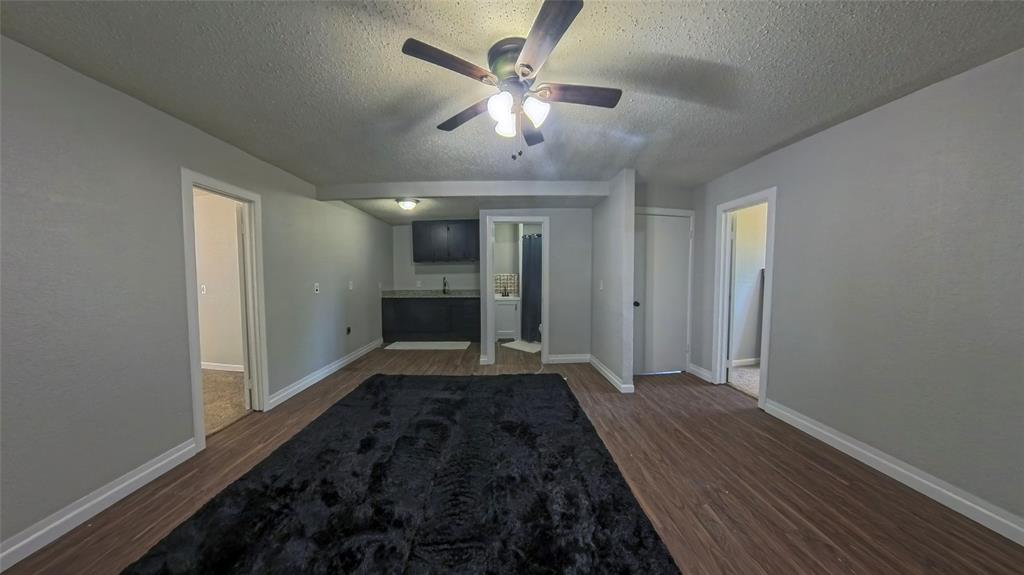 Room featuring wood-finish flooring, a ceiling fan with integrated lighting, a kitchenette with dark cabinetry, and neutral wall tones