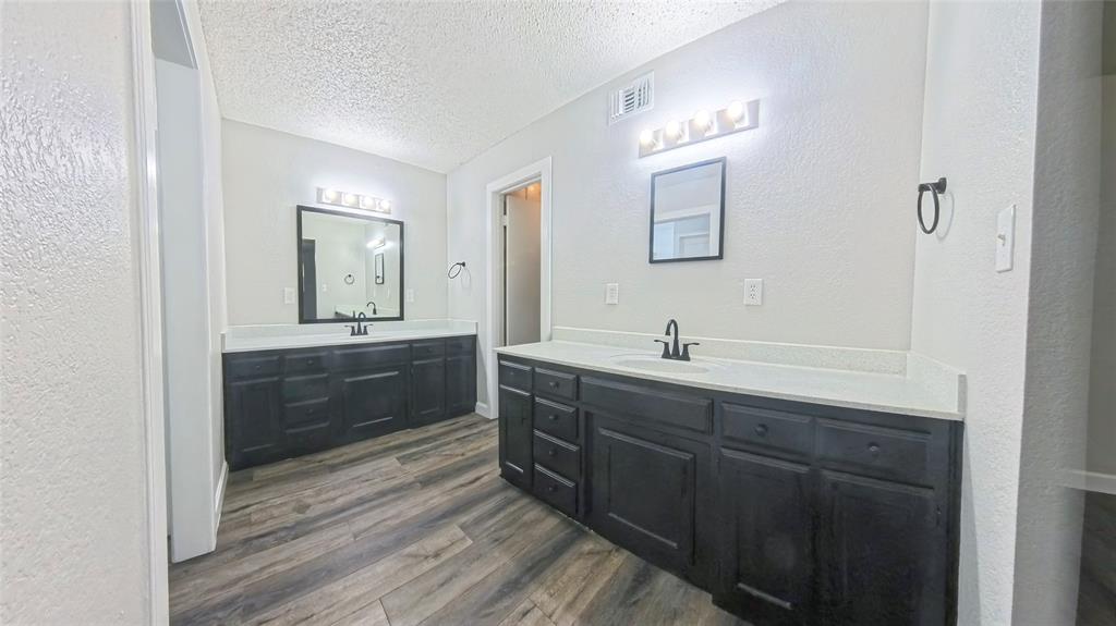 Dual-sink vanity featuring dark-toned cabinetry, light-toned countertops, wood-finish flooring, and wall-mounted vanity lighting