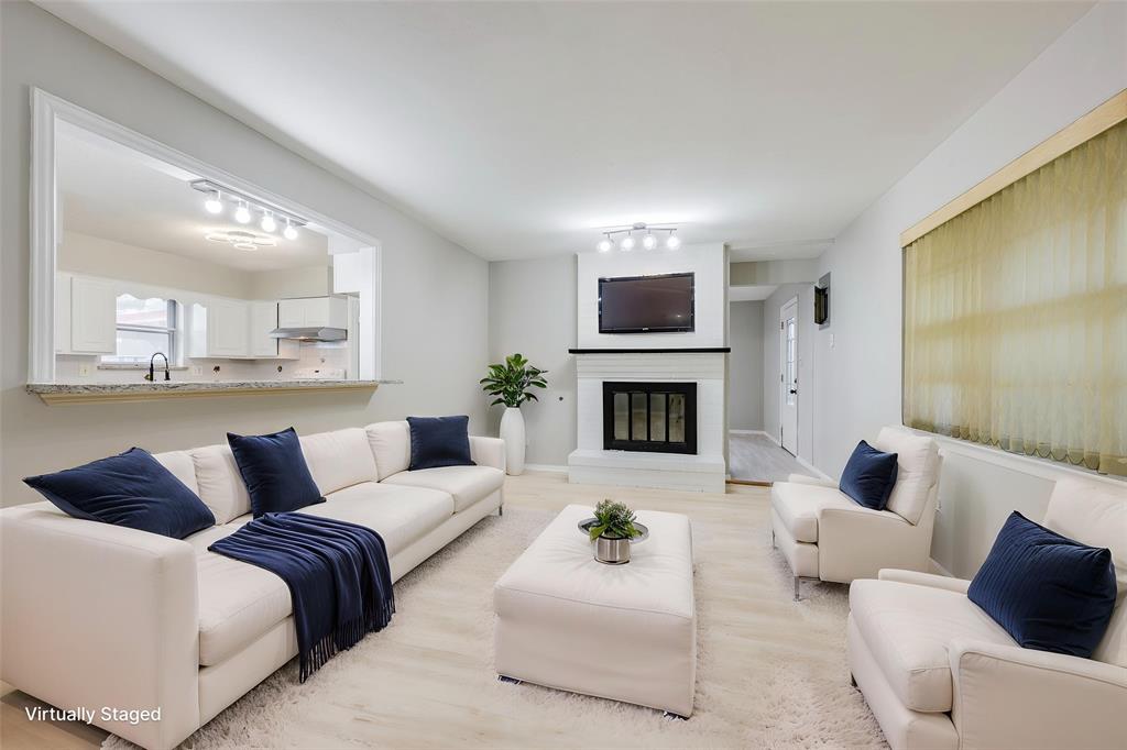 Living area featuring light-toned wood-finish flooring, a painted brick fireplace with a dark mantel, a pass-through opening to the kitchen, and modern multi-bulb track lighting