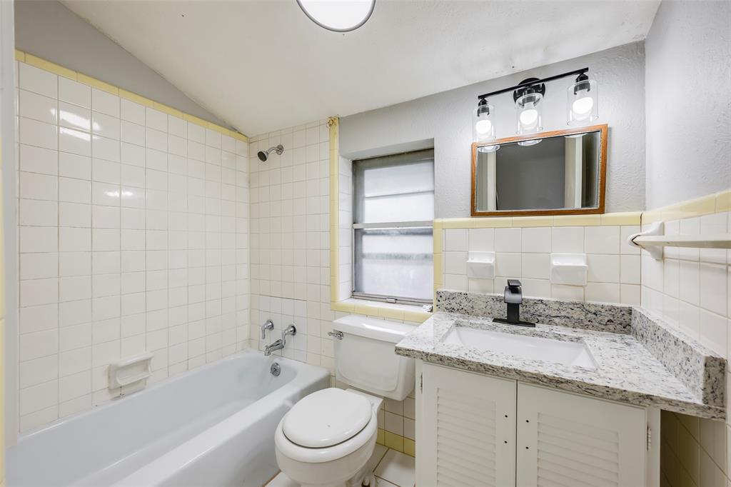 Bathroom featuring a vanity with a stone-finish countertop, white louvered cabinetry, wall-mounted three-light fixture, and tile-surround tub