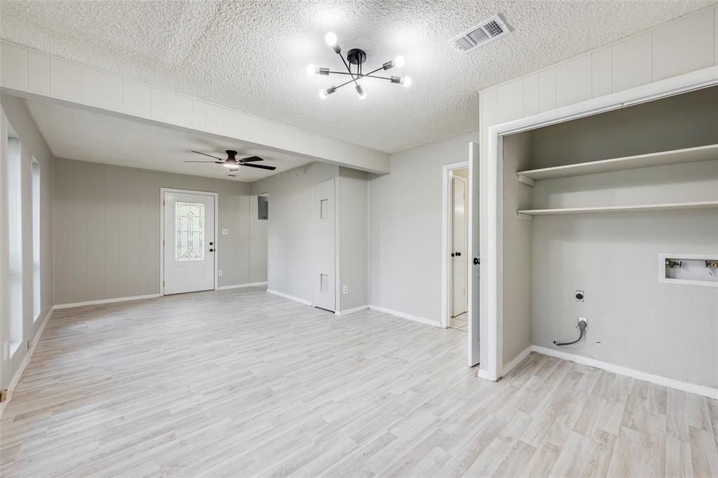 Open-concept interior featuring light wood-finish flooring, a multi-arm modern ceiling fixture, built-in wall shelving, and an exterior door with decorative glass paneling