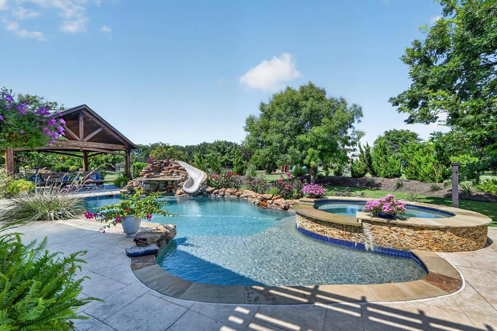 Custom swimming pool featuring a rock waterfall, integrated slide, elevated stone-clad spa, and sprawling stamped concrete patio with a detached timber-frame pavilion