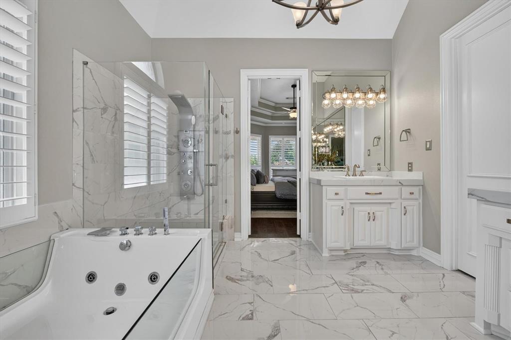 Bathroom featuring large-format tile flooring, jetted soaking tub, glass-enclosed shower with body spray system, white cabinetry with stone countertops, and a wall-to-wall vanity mirror with integrated lighting