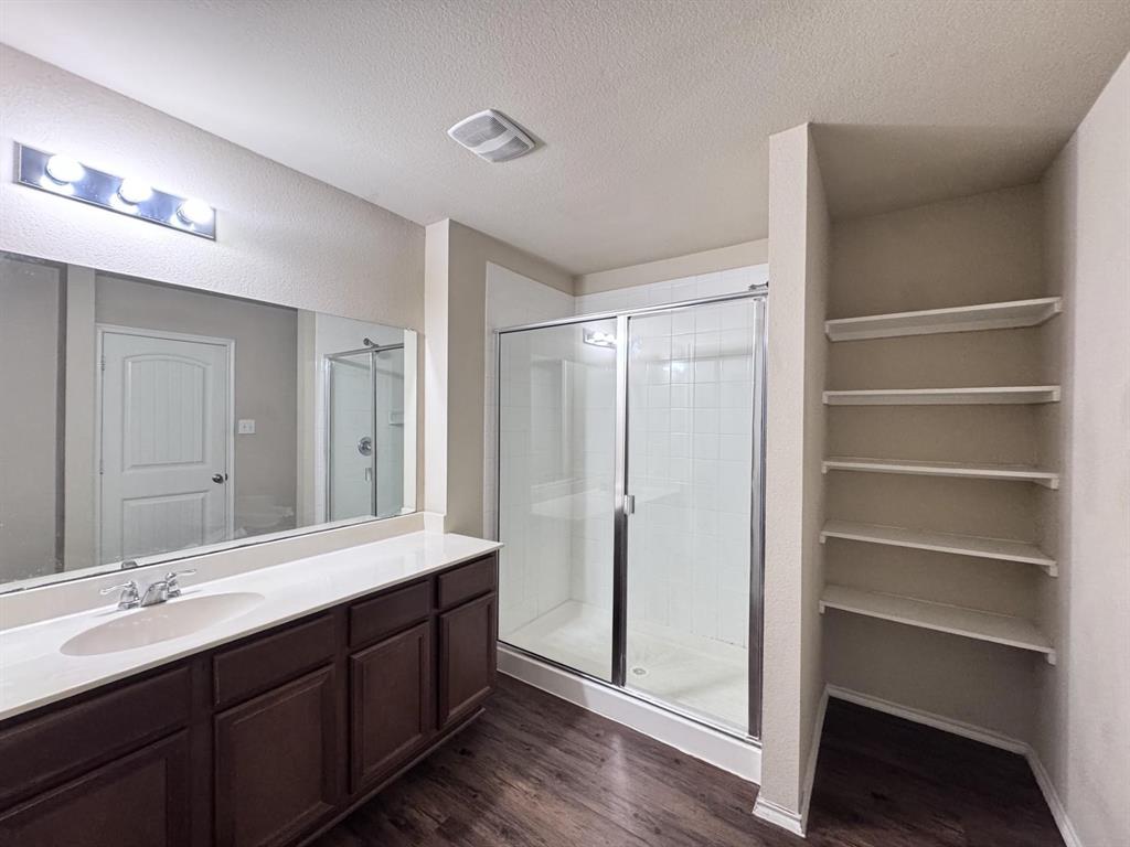 Bathroom featuring a vanity with a white countertop and oval sink, a glass-enclosed shower, wood-finish flooring, a full-width vanity mirror, and built-in wall shelving